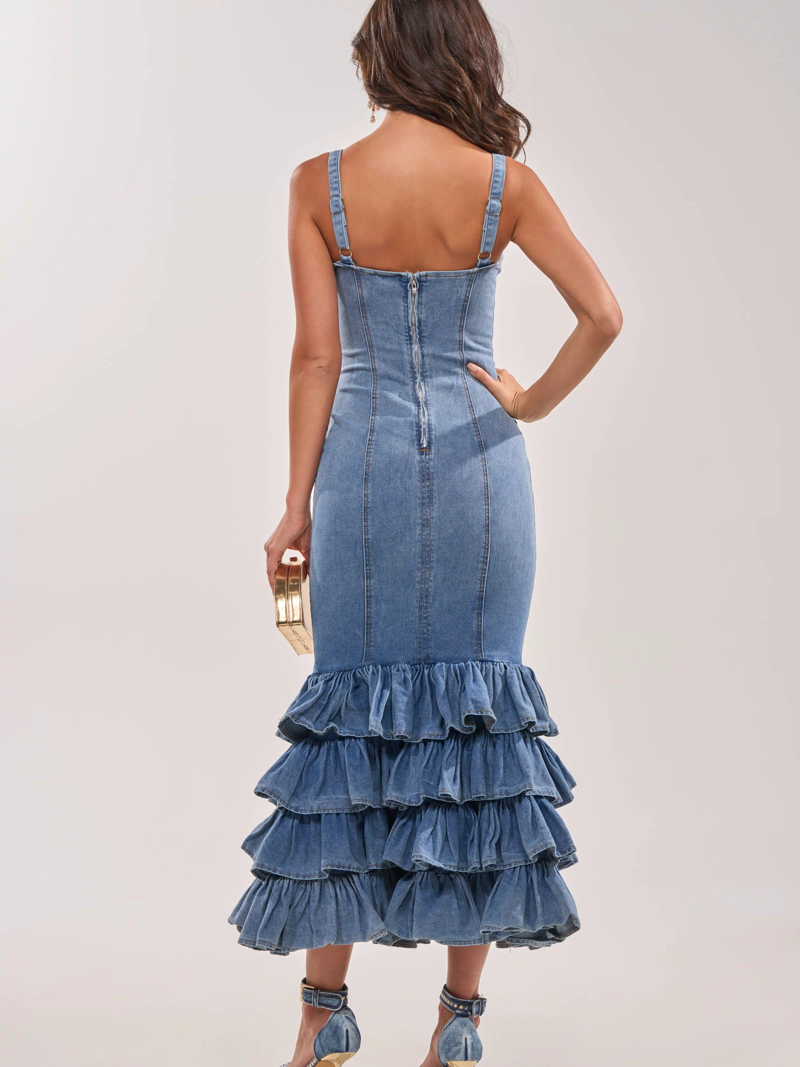 A woman faces away wearing the MAIN ONE DENIM RUFFLE MAXI DRESS with a tiered ruffle hem. She holds a gold clutch and wears strappy high-heeled sandals against a plain light-colored background.