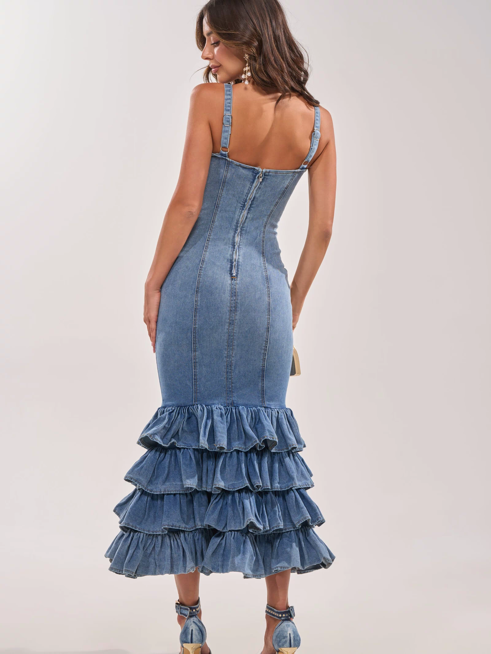 A woman faces away wearing the MAIN ONE DENIM RUFFLE MAXI DRESS featuring thin straps, a back zipper, and a tiered ruffle hem. She pairs it with heeled sandals and carries a small purse.