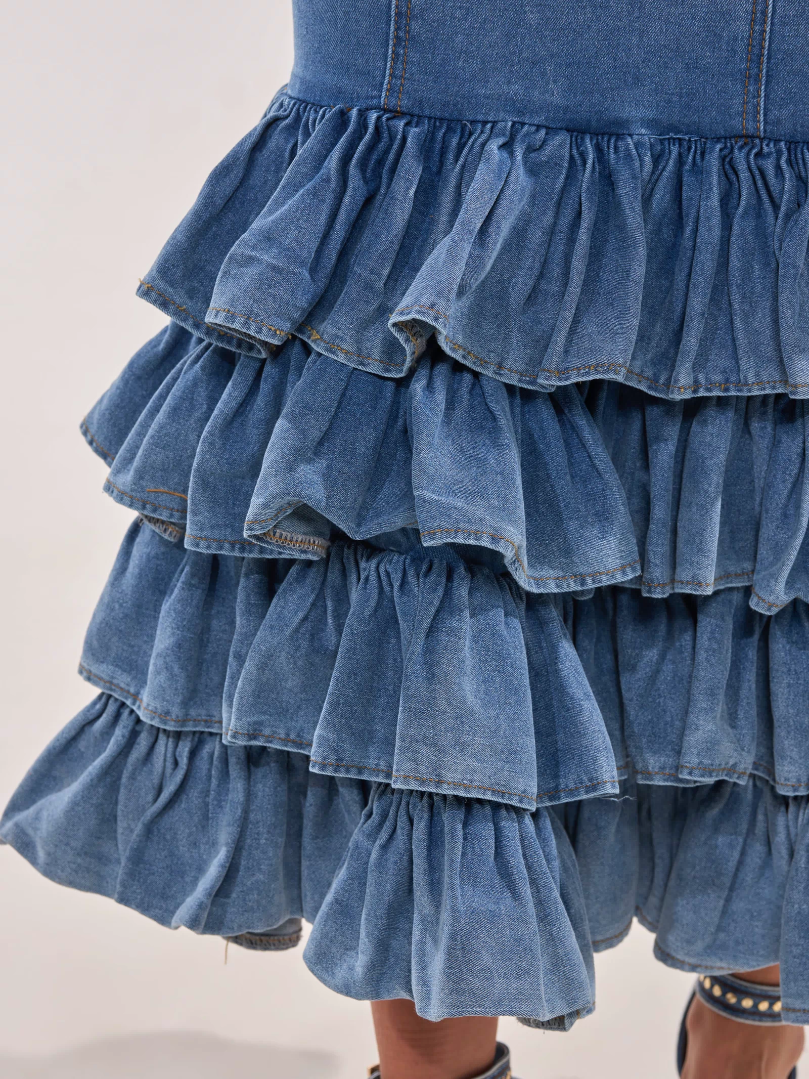 Close-up of the MAIN ONE DENIM RUFFLE MAXI DRESS, featuring tiered ruffled blue denim layers reminiscent of mermaid-inspired style. The hem is above the knee, paired with a sandal with denim straps partially visible below.