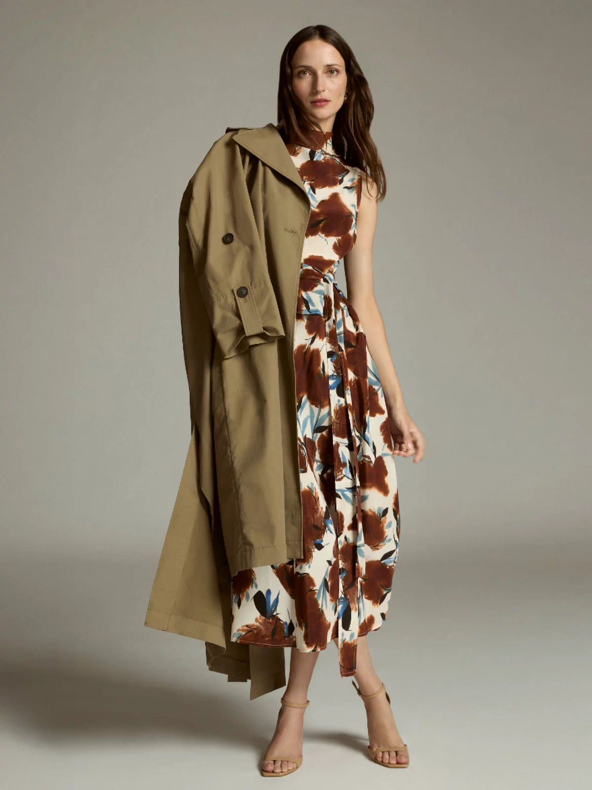 Woman wearing a patterned dress with a trench coat draped over her shoulder against a neutral background
