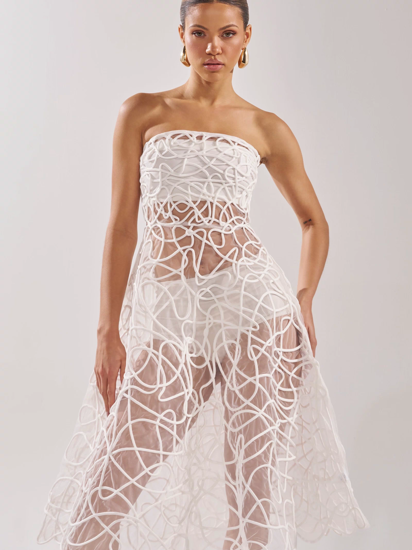 A woman models the MAYA ABSTRACT MIDI DRESS IN WHITE, a strapless sheer midi dress with an abstract white pattern—an elegant cocktail party choice—while standing against a plain light background.