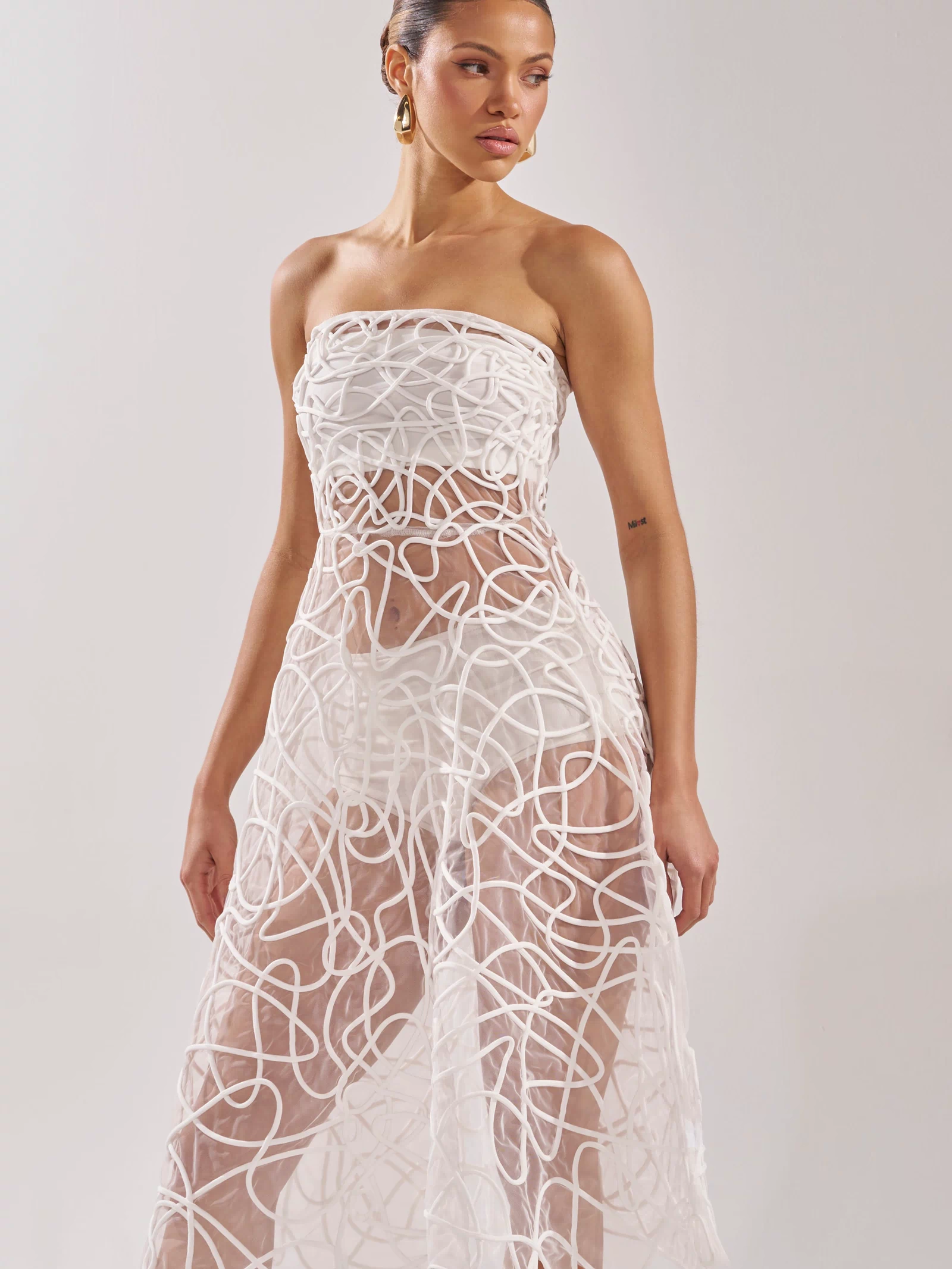 She wears the MAYA ABSTRACT MIDI DRESS IN WHITE, a strapless sheer midi with swirling textured lines and a flattering fitted bodice. Her hair is pulled back and she pairs gold earrings, posing against a plain light background.