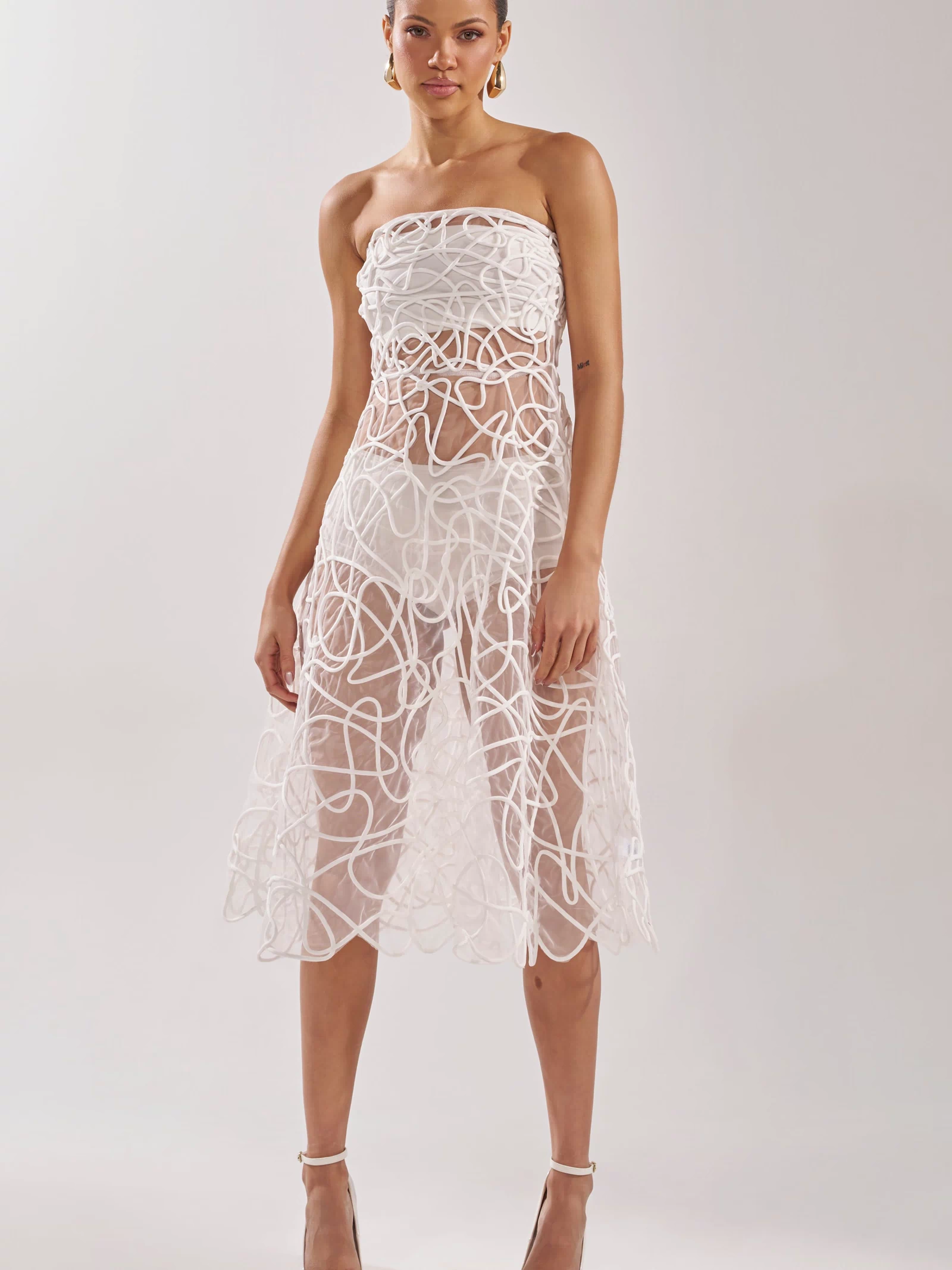 A woman wears the MAYA ABSTRACT MIDI DRESS IN WHITE—a strapless sheer midi with swirling patterns—styled with gold pointed-toe heels and hoop earrings, standing against a plain background, perfect for cocktail parties.