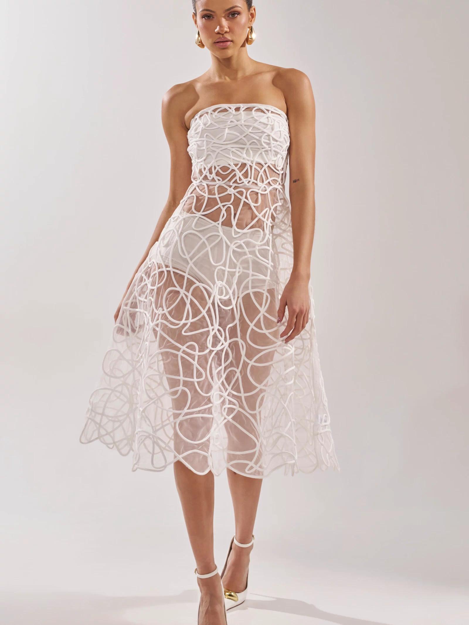 A woman wears the MAYA ABSTRACT MIDI DRESS IN WHITE, a strapless sheer dress with a geometric swirl overlay, fitted bodice, and mid-calf skirt. Gold hoop earrings and pointed-toe heels complete her chic cocktail party look.