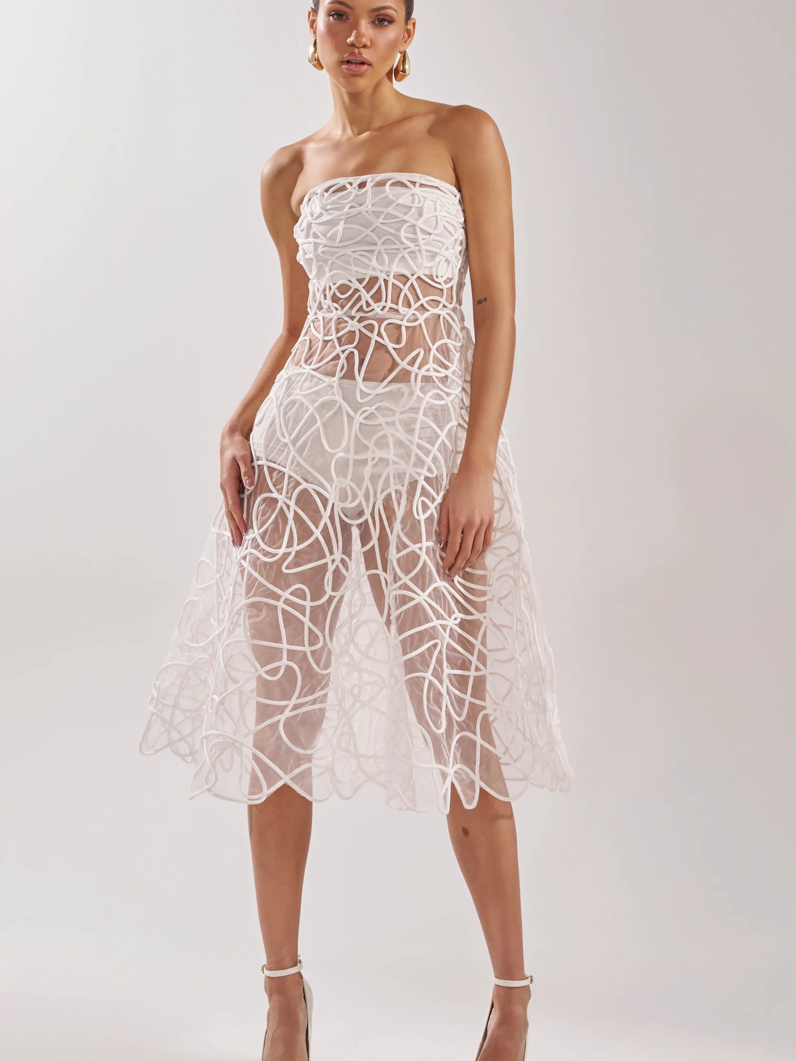 A woman poses against a plain background in the MAYA ABSTRACT MIDI DRESS IN WHITE, a strapless sheer midi dress with bold swirling white patterns. She completes the look with white high heels and gold hoop earrings.