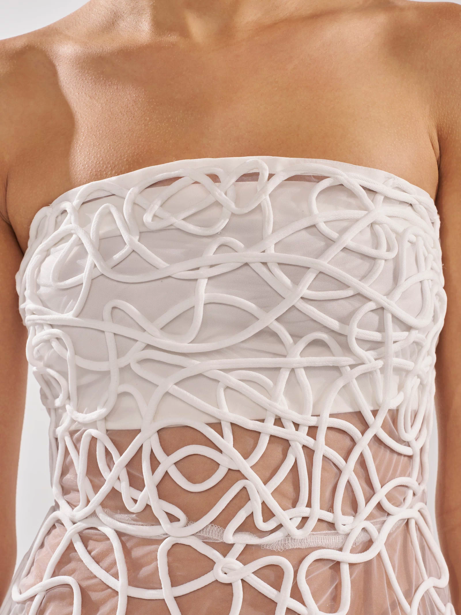 A close-up of a person wearing the MAYA ABSTRACT MIDI DRESS IN WHITE, a strapless sheer evening dress with textured, three-dimensional white lines forming an abstract, embroidered swirling pattern over sheer fabric.