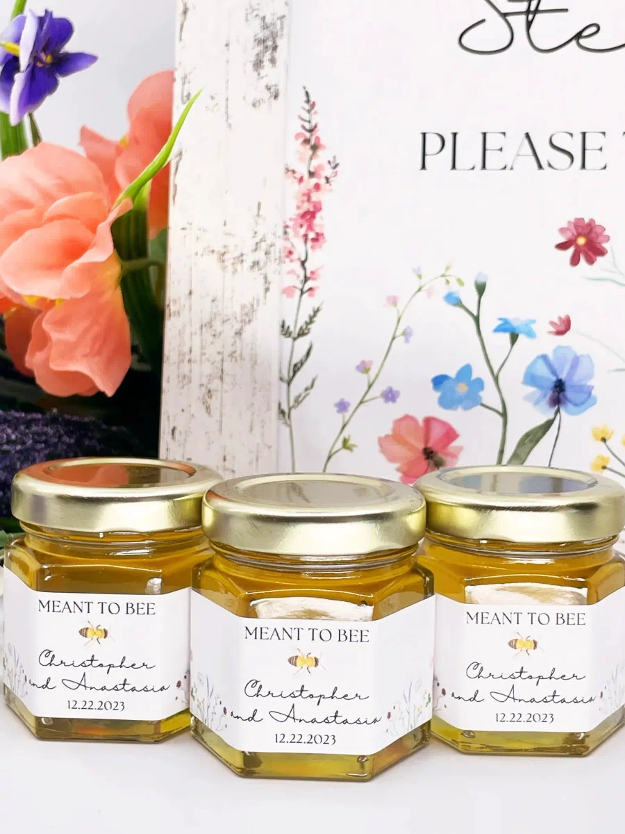 Three jars of honey with 'Meant to Bee' labels on a white surface, surrounded by flowers.