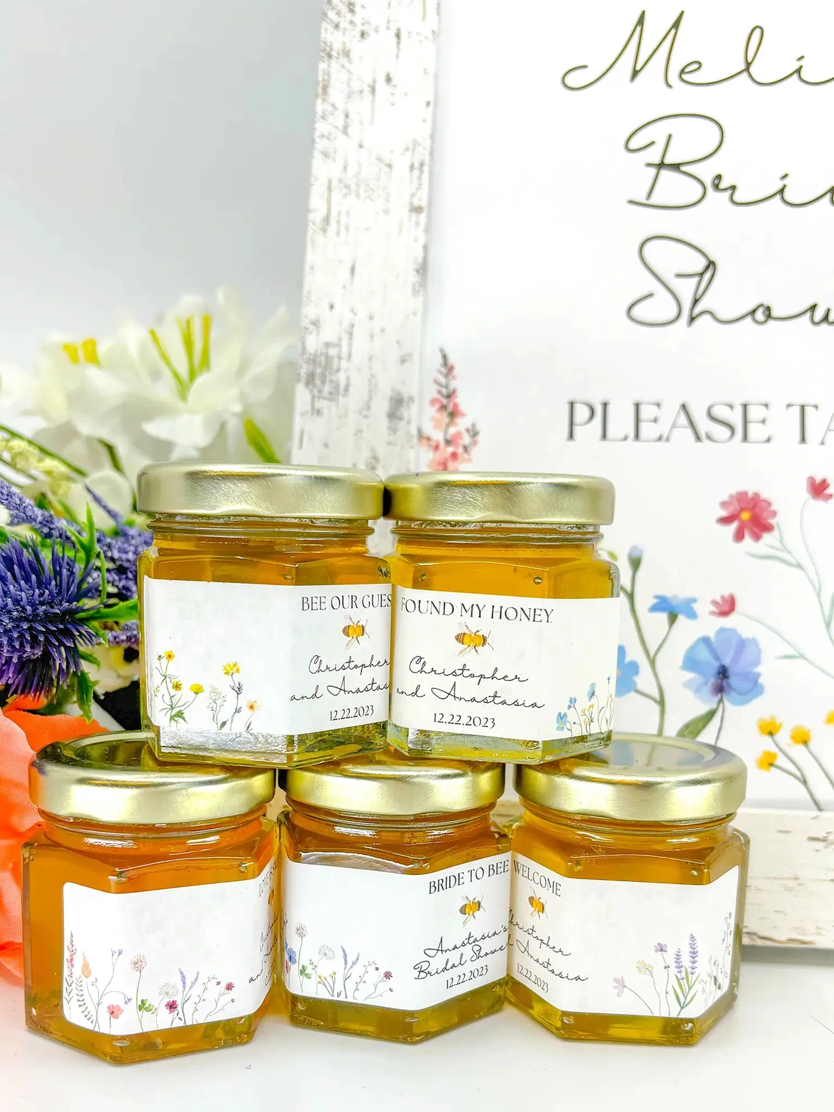 Small jars of honey with personalized labels, honey, and gold lids. Title options include Bee Our Guest, Found My Honey, Bride to Bee, Welcome, Love is Sweet, and more. Showcasing different flower color options: multi, yellow, blue, red/pink, and purple.