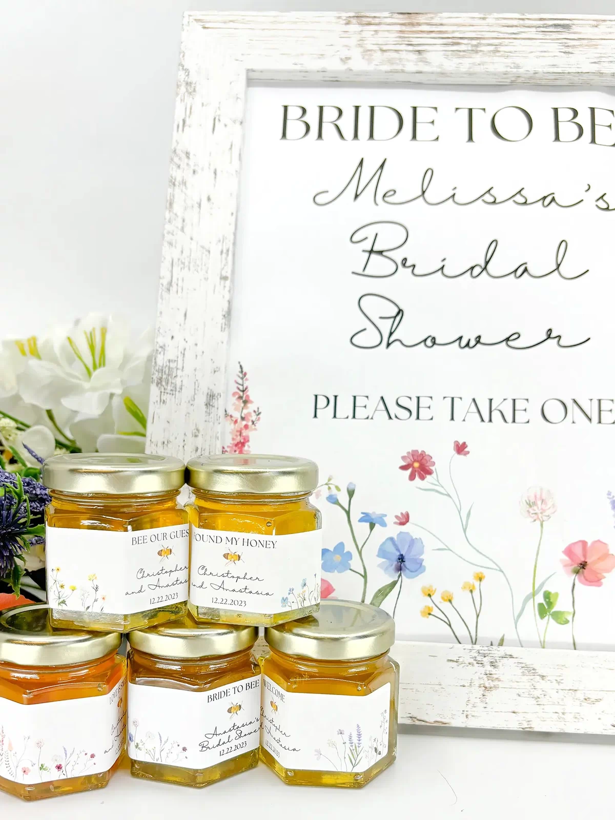 Small jars of honey with personalized labels and gold lids in front of bridal shower sign. Label titles include Bee Our Guest, Bride to Bee, Found My Honey, Welcome, Love is Sweet, and more. Floral color options shown: red/pink, purple, yellow, blue, and multi. 