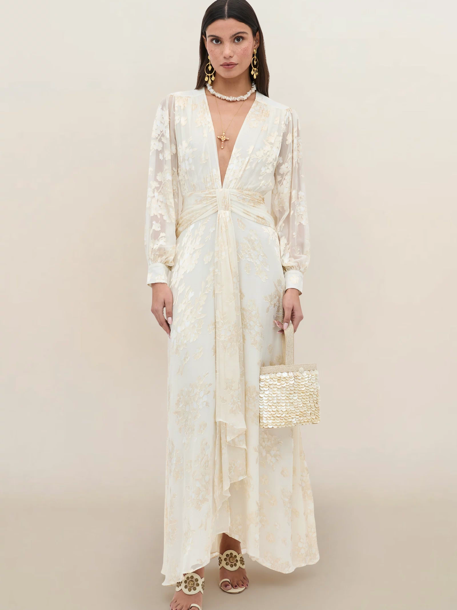 A woman wears the Meera - Bouquet Devoré Cream gown, a long cream silk-satin dress with floral patterns, deep V-neckline, long sleeves, and draped front. She complements it with a woven handbag, gold earrings, layered necklaces, and heeled sandals.