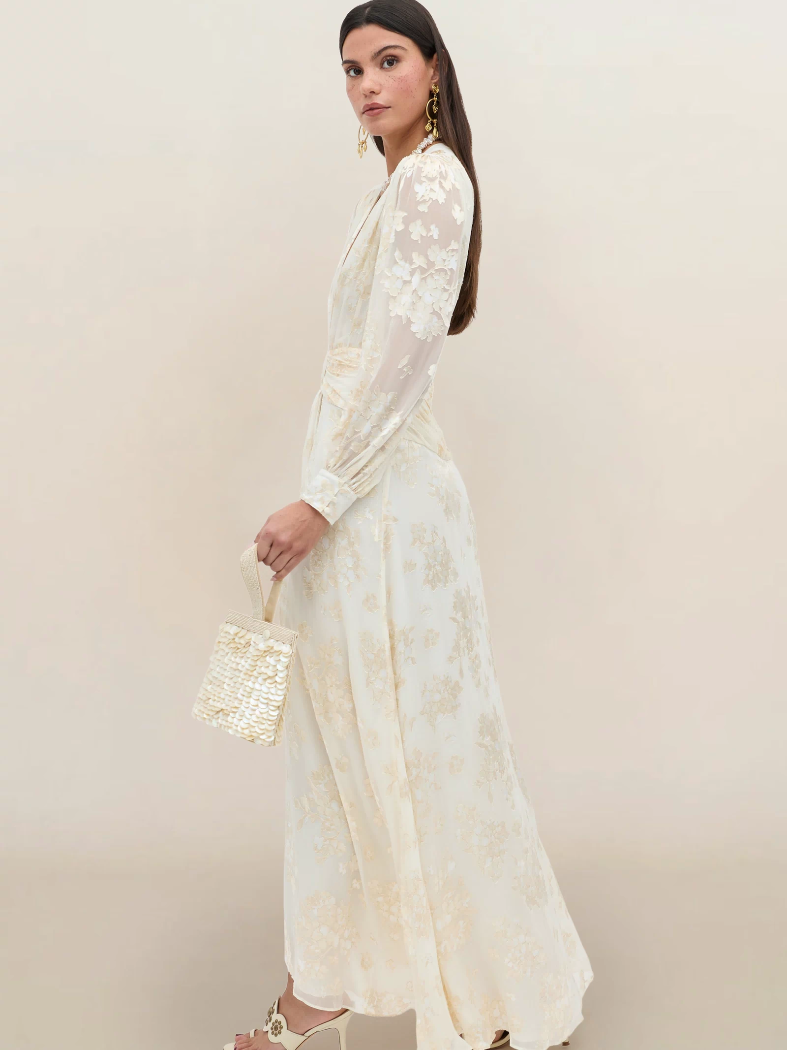 A woman stands sideways in the Meera - Bouquet Devoré Cream, a long cream silk-satin gown with floral details and sheer sleeves. She holds a woven handbag, wears heeled sandals and gold earrings, posing before a plain beige backdrop.