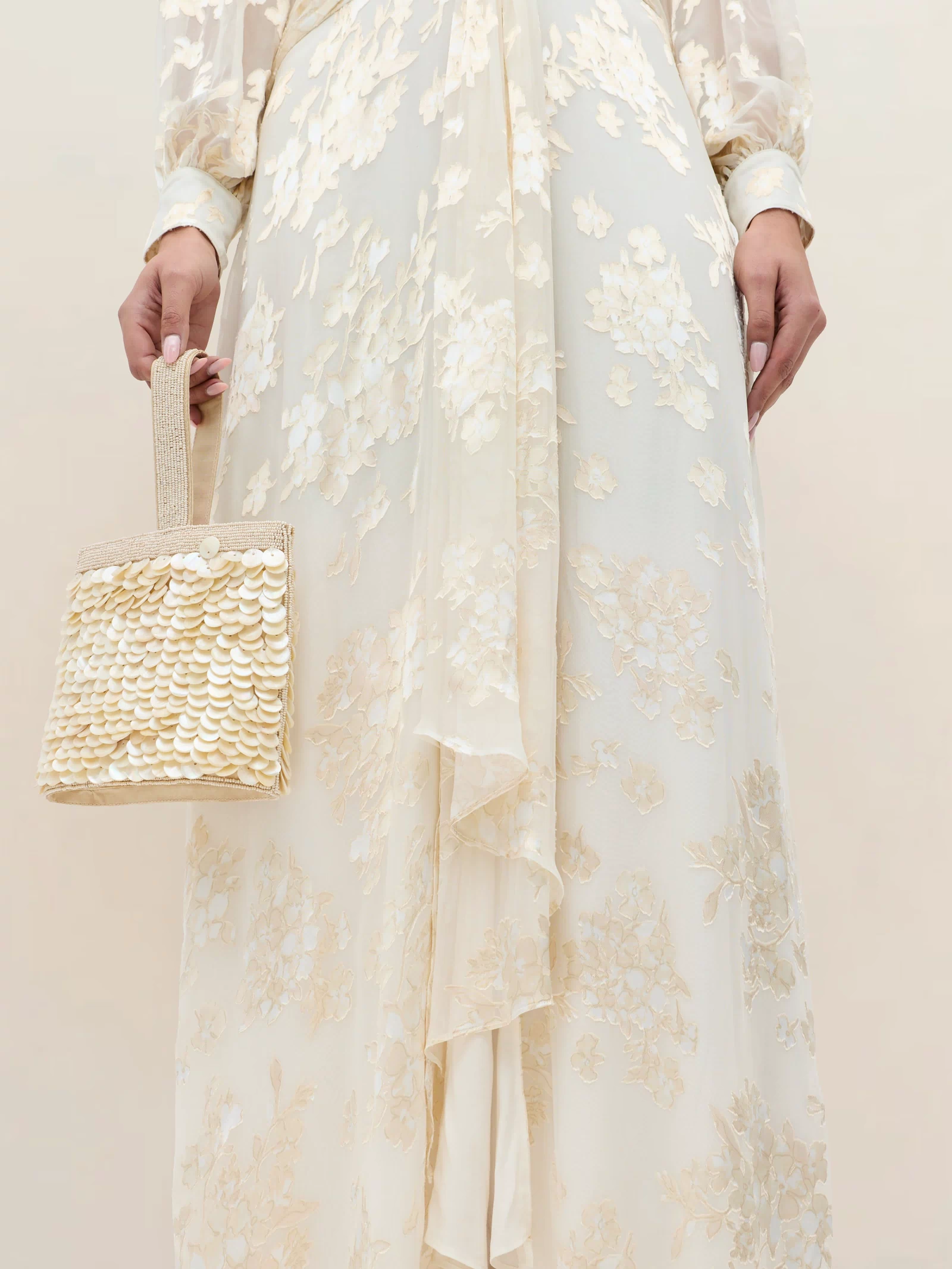 A person wears the Meera - Bouquet Devoré Cream, a long cream silk-satin gown with floral embroidery, holding a small textured beige bag with sequin-like embellishments. The photo shows the outfit from shoulders to knees.