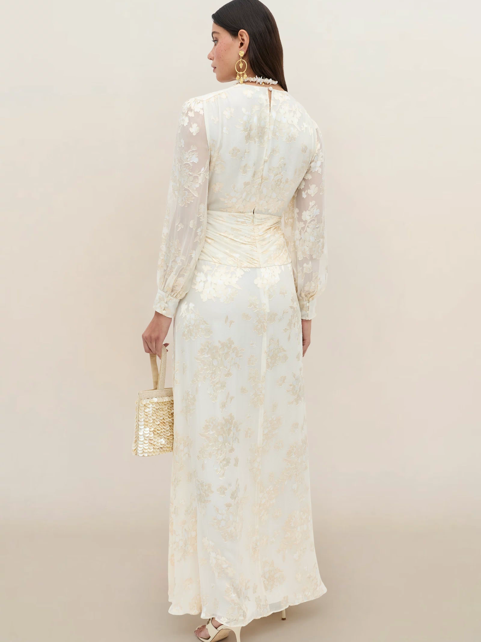 A woman faces away in the Meera - Bouquet Devoré Cream silk-satin gown with floral details and sheer long sleeves, holding a woven bag. She wears gold earrings and has shoulder-length dark hair against a light background.