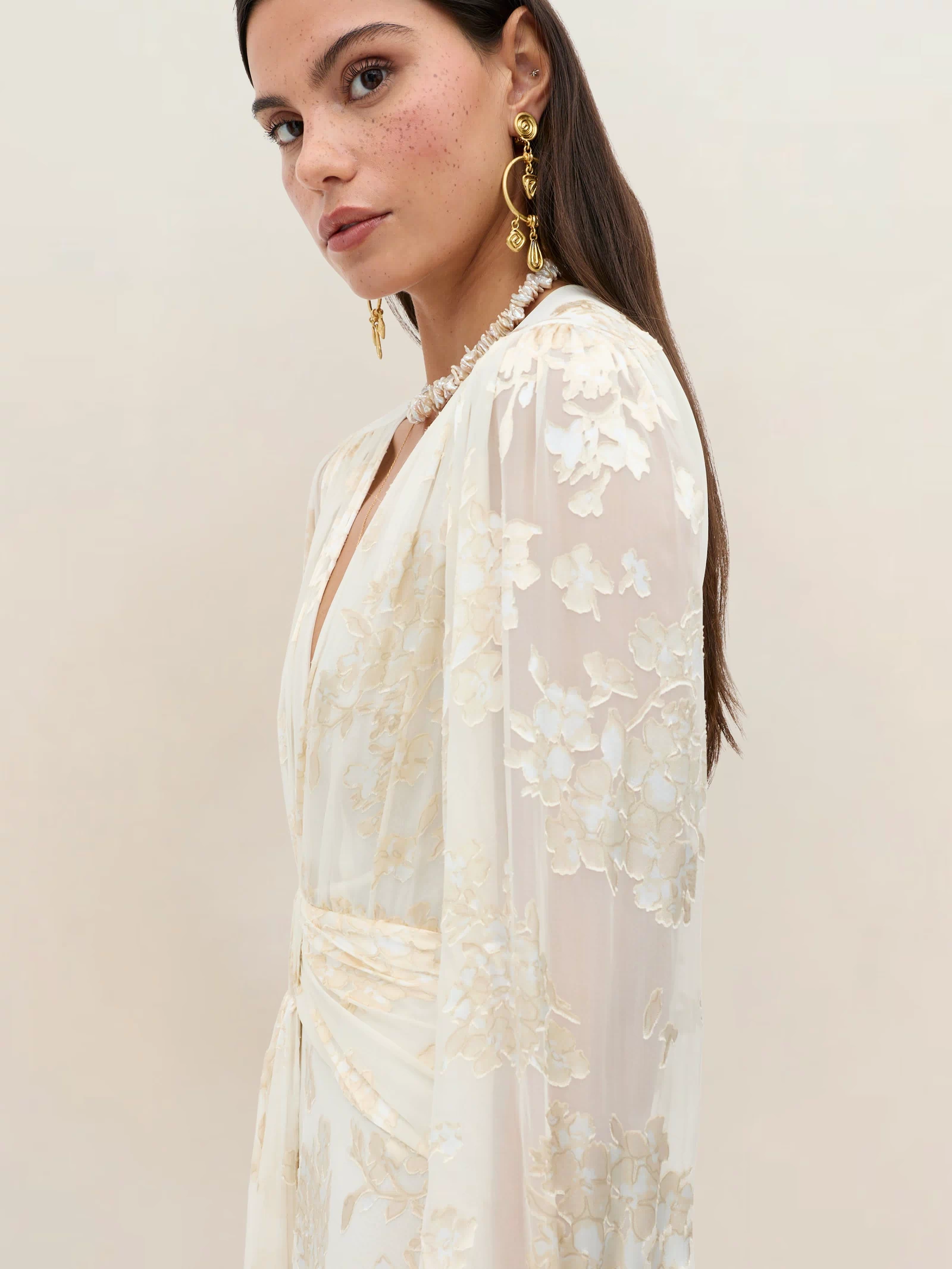A woman with long brown hair and freckles models the Meera - Bouquet Devoré Cream bridal gown, featuring sheer cream floral fabric and long sleeves, paired with gold statement earrings against a plain light background.
