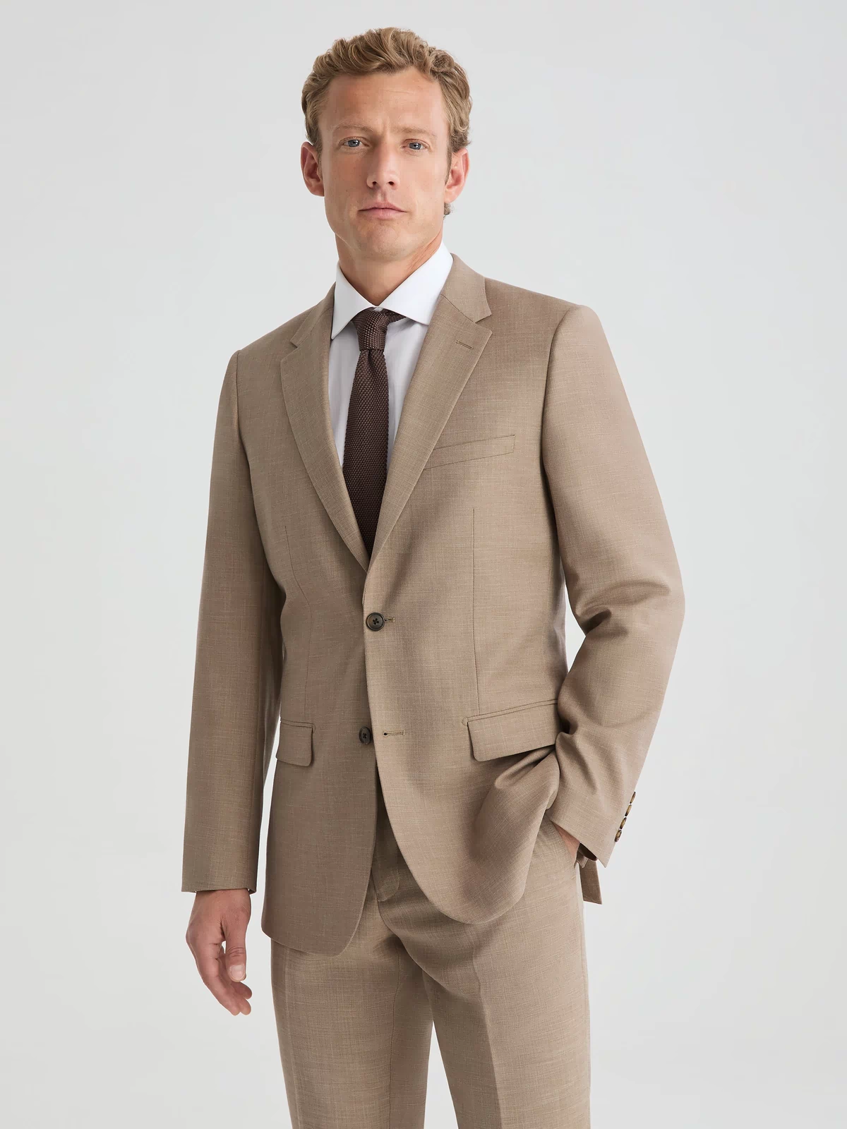 Man wearing dark tan suit jacket and suit pants. Paired with white long sleeve button up shirt and dark brown tie.