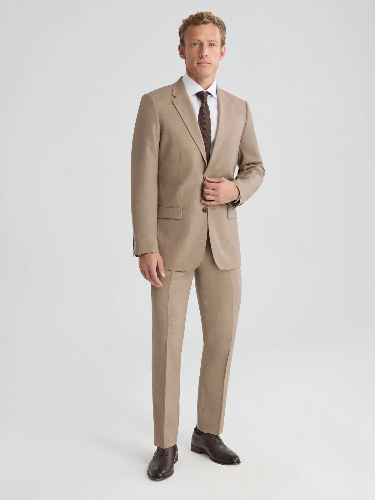 Man wearing dark tan suit jacket and suit pants. Paired with white long sleeve button up shirt and dark brown tie.