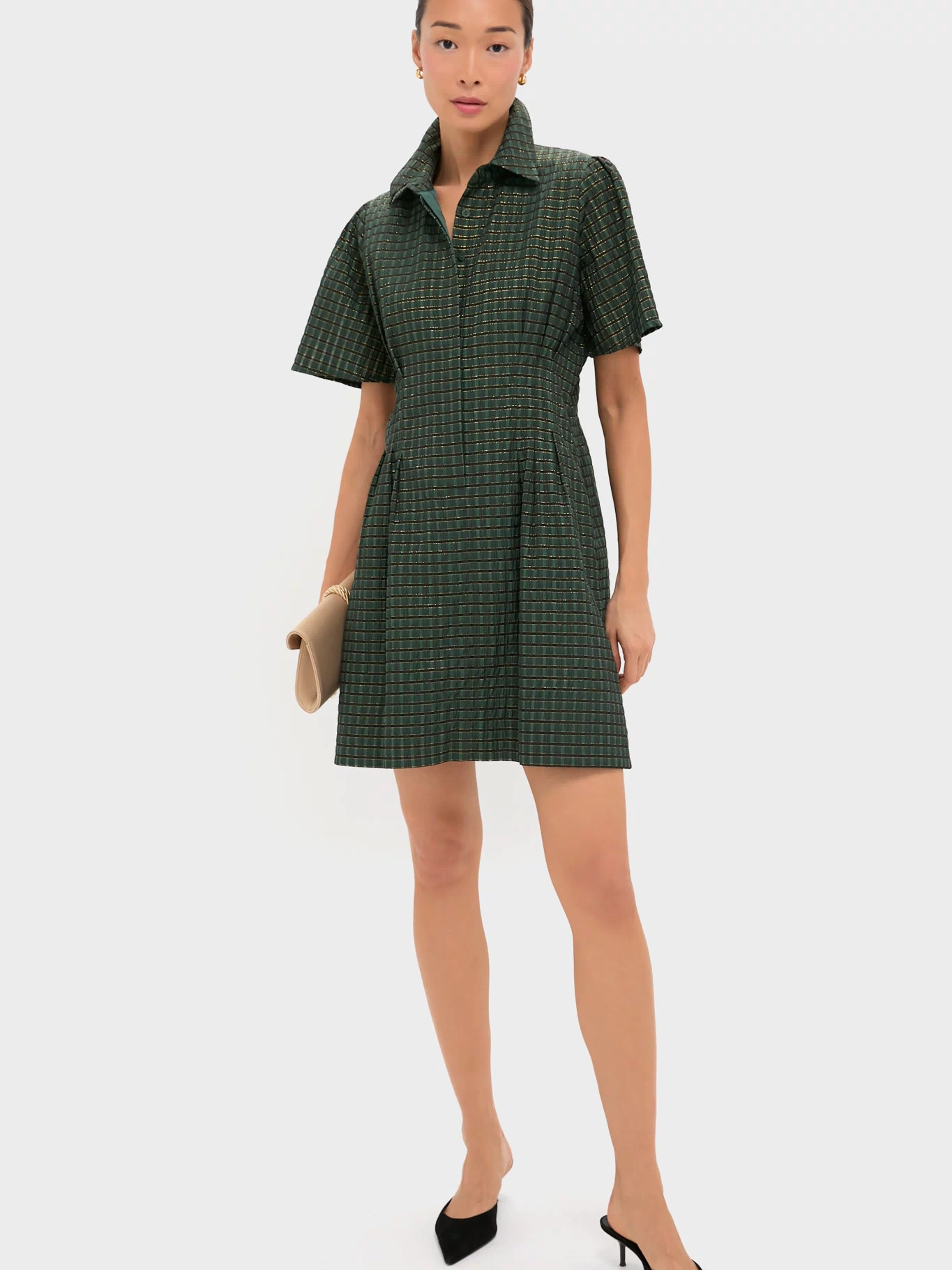 A woman wears the Tuckernuck Metallic Green Plaid Mini Delaney Dress—featuring short sleeves, a collar, and a subtle check pattern—styled with black pointed heels and a beige clutch.