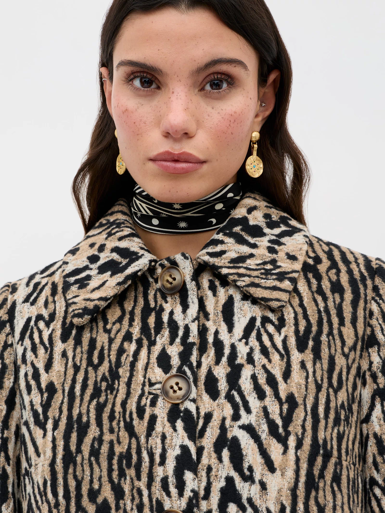 A woman with long dark hair and freckles wears the Milly - Bohemia Leopard mid-length coat with large buttons, gold drop earrings, and a black-and-white scarf around her neck, standing against a plain light background.