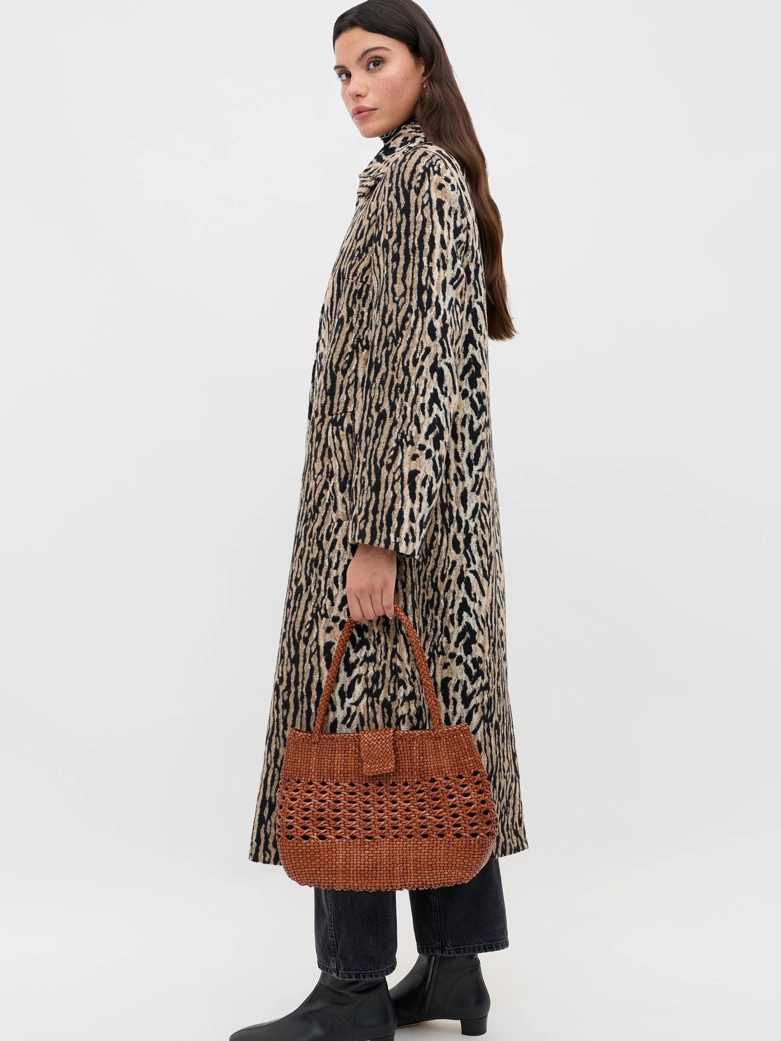 A woman wears the Milly - Bohemia Leopard mid-length coat, paired with black pants and boots, holding a brown woven handbag as she stands against a plain white background and glances over her shoulder.
