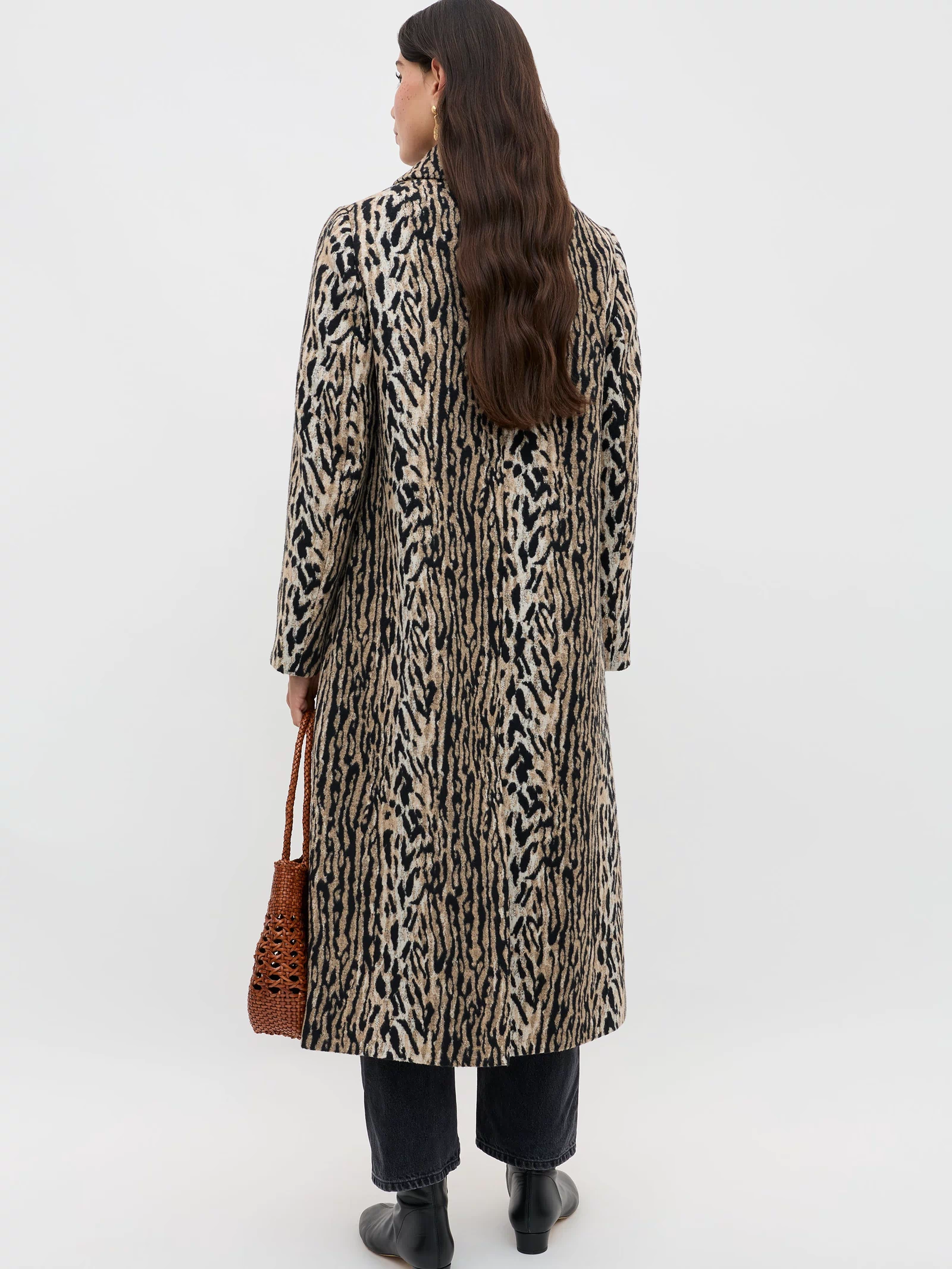 A person with long brown hair stands facing away, wearing the Milly - Bohemia Leopard mid-length coat, black pants and boots, holding a brown woven bag against a plain light background.