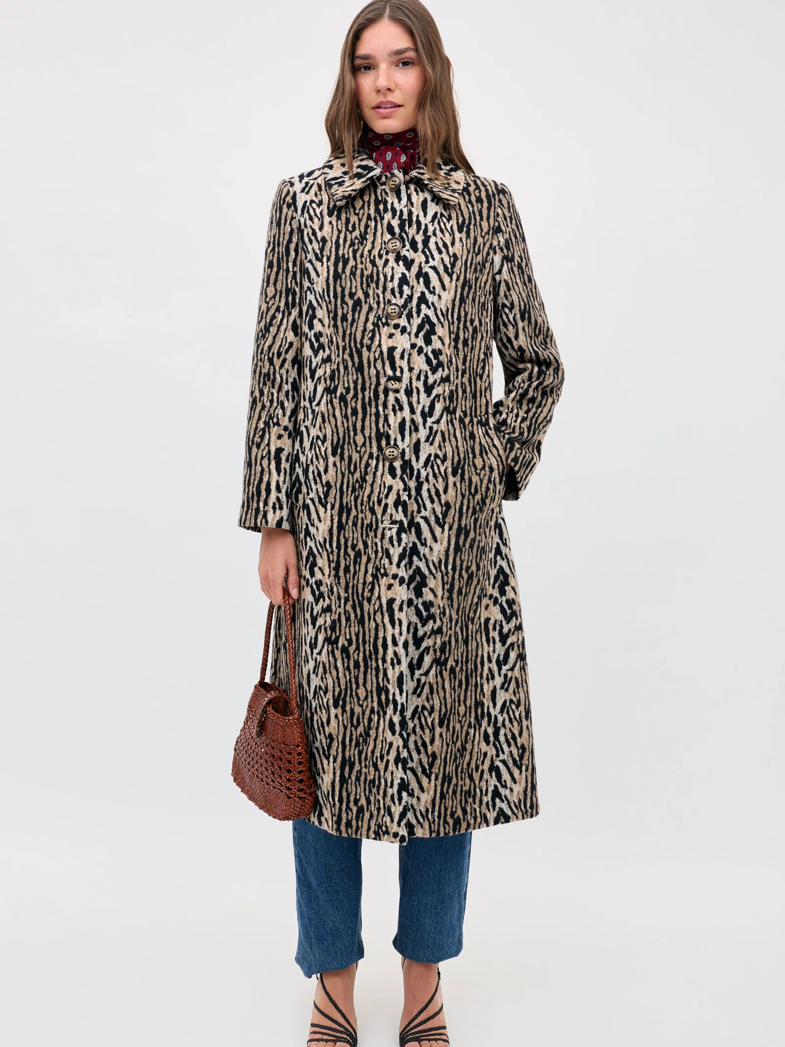 A woman in the Milly - Bohemia Leopard coat over blue jeans and black strappy sandals holds a brown woven handbag, facing the camera against a plain white background.