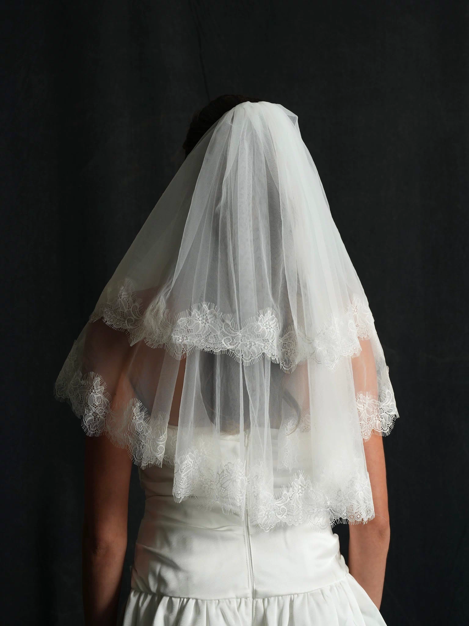 Person wearing a white wedding dress with the back profile of a mini chantilly lace short two tier bachelorette veil 