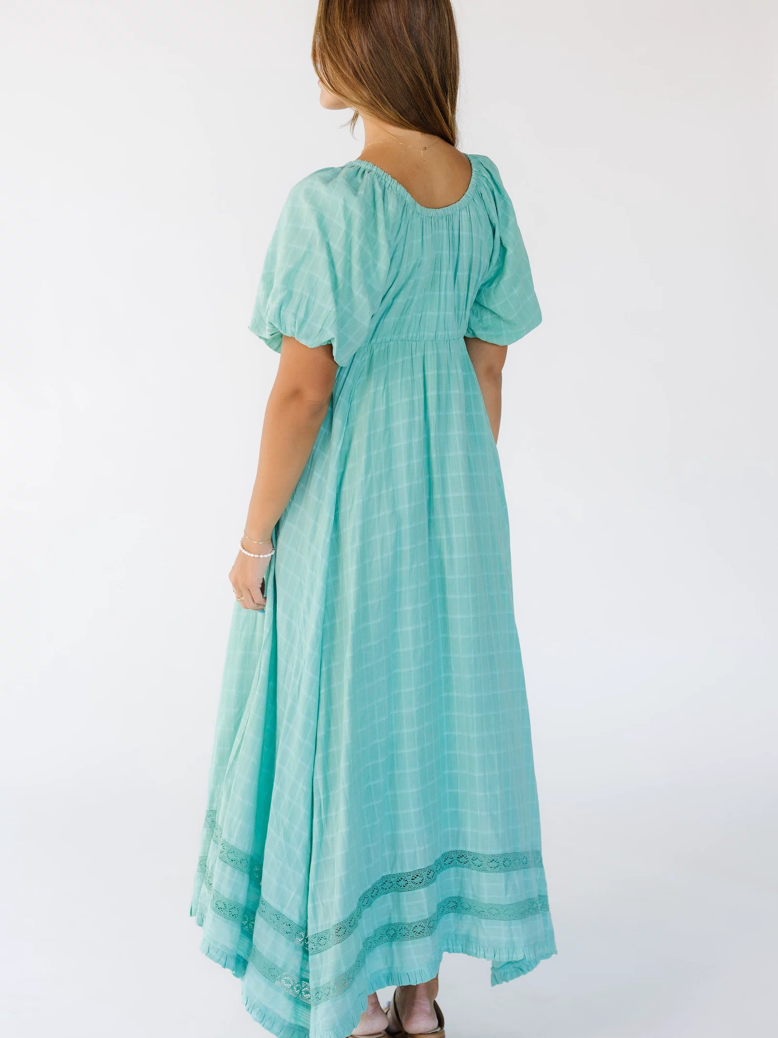 Back view of a young woman wearing a mint green, loose fitting midi-maxi dress with lace insets and a scoop back neckline.