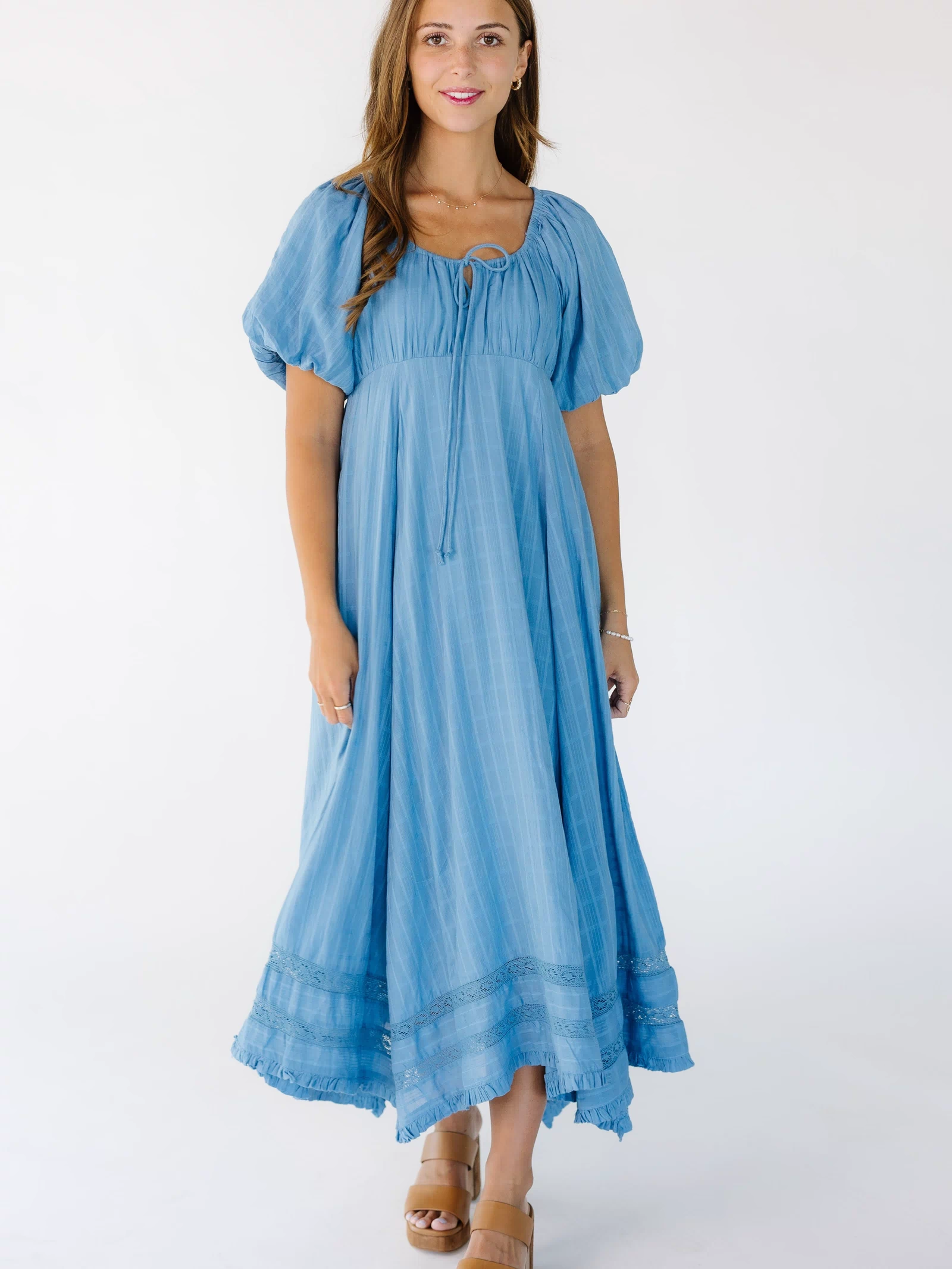 Woman wearing a blue midi-maxi dress with 2 rows of lace inset near the narrow ruffle hem with a scoop neckline and balloon sleeves.