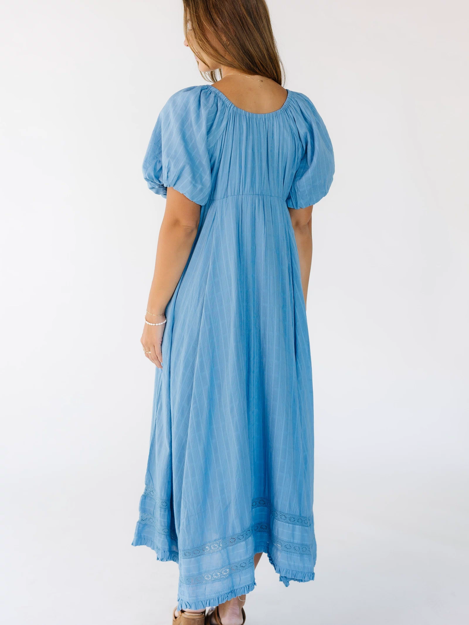 Back view of a young woman wearing a blue midi-maxi dress with balloon sleeves and a scoop back neckline.