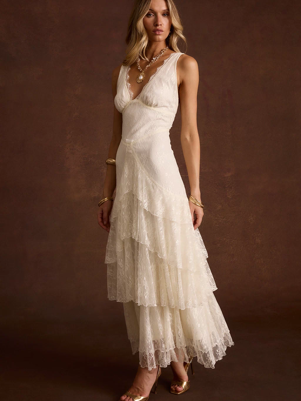 Cream lace maxi dress with V-neck and tiered ruffle skirt, styled with gold heels.