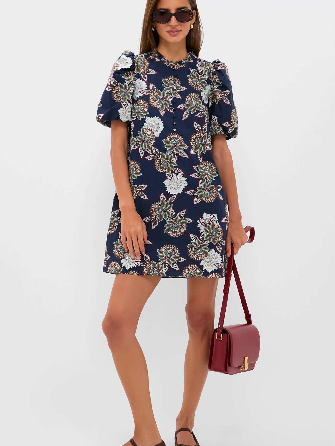 Woman in Navy Floral Mimi Mini Dress with puff sleeves, accessorized with dark sunglasses, gold hoop earrings, a burgundy crossbody bag, and brown ballet flats, exuding classic, effortless style.