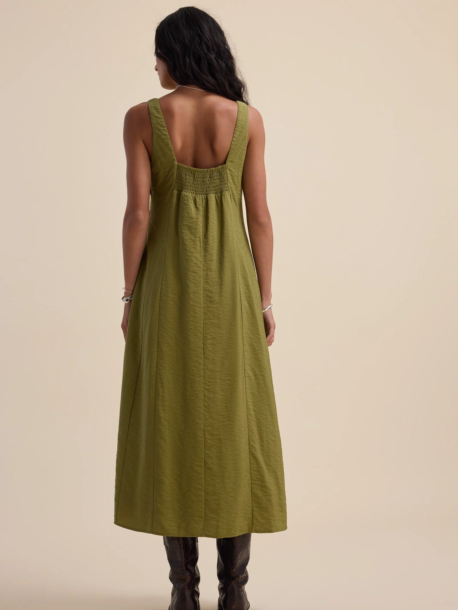 Woman in a Nina Fit and Flare Dress by Max, dill green, sleeveless, with brown boots, rear view.