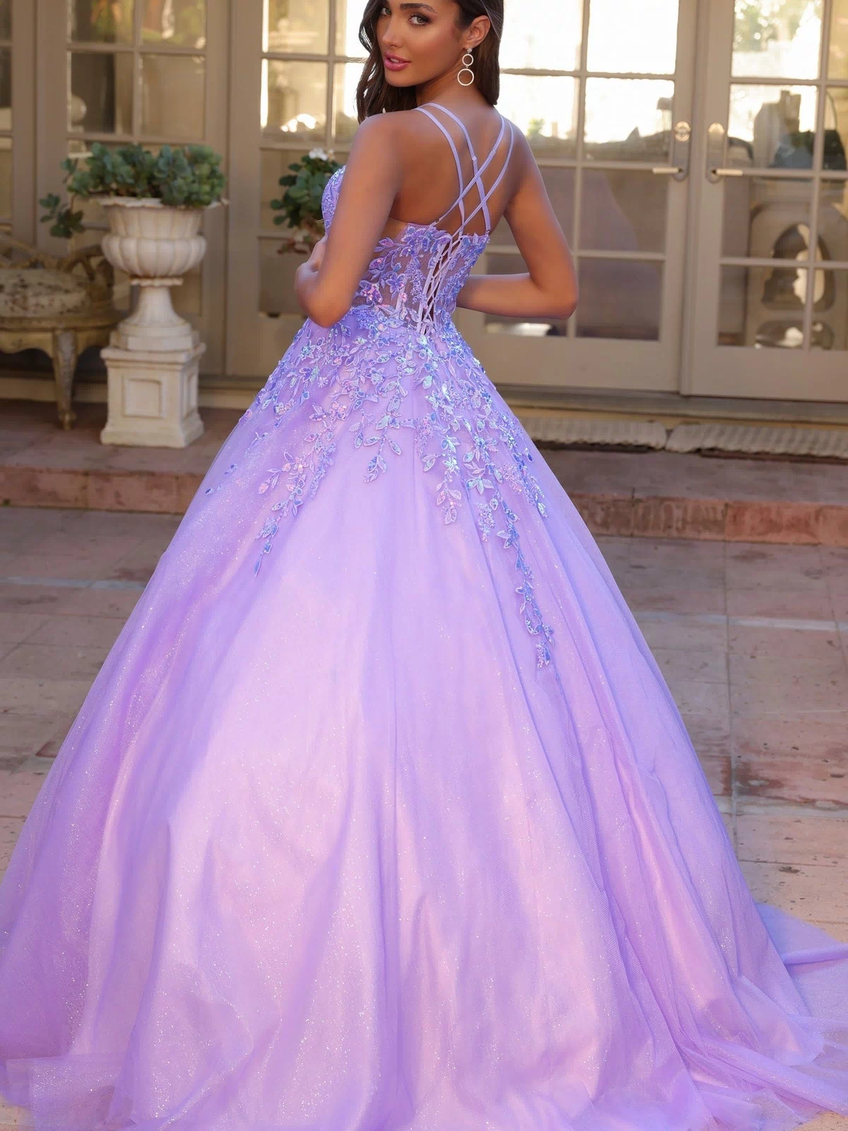 A woman in the Glitter-Tulle Long Prom Ball Gown H1271 with floral accents and a strappy back stands on a patio, looking over her shoulder.