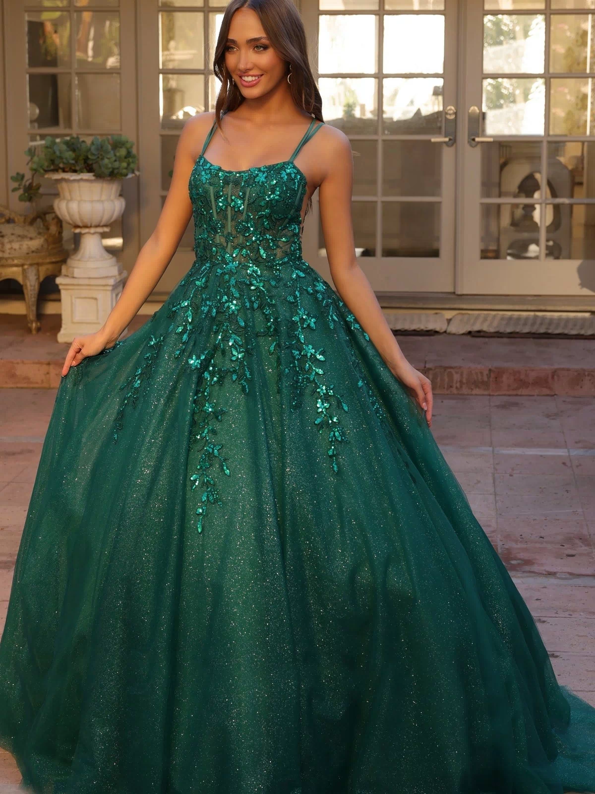 A woman stands outside in front of glass doors, wearing the Glitter-Tulle Long Prom Ball Gown H1271, a shimmering emerald green dress with floral embellishments and thin straps.