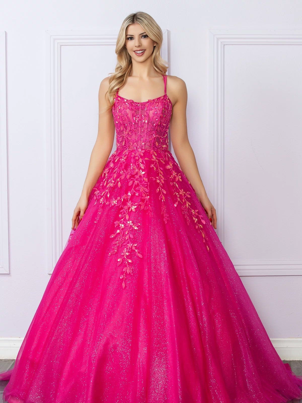 A woman in a Glitter-Tulle Long Prom Ball Gown H1271, featuring bright pink color and floral embellishments, stands before a light-colored paneled wall.