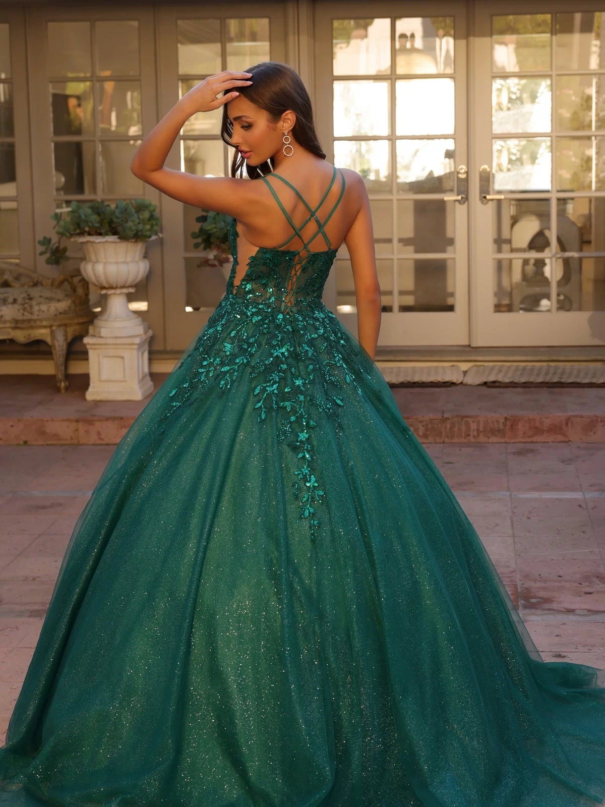 A woman stands outside in front of glass doors, wearing the Glitter-Tulle Long Prom Ball Gown H1271 with floral details and crisscross back straps.