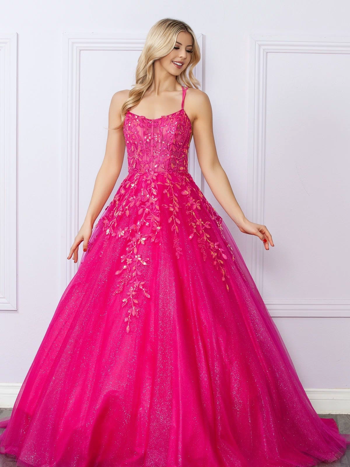 A woman stands against a light wall, smiling to the side in the Glitter-Tulle Long Prom Ball Gown H1271—a bright pink, floor-length dress with floral embellishments.