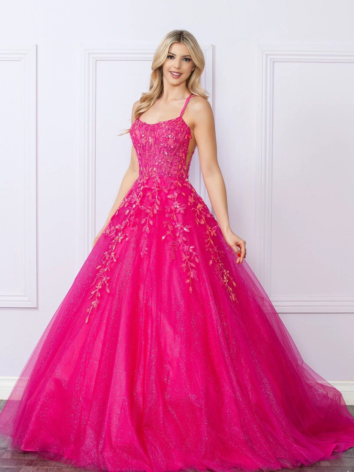 A woman poses indoors by a light-colored, white-paneled wall wearing the Glitter-Tulle Long Prom Ball Gown H1271, a bright pink dress with thin straps and delicate floral embroidery.
