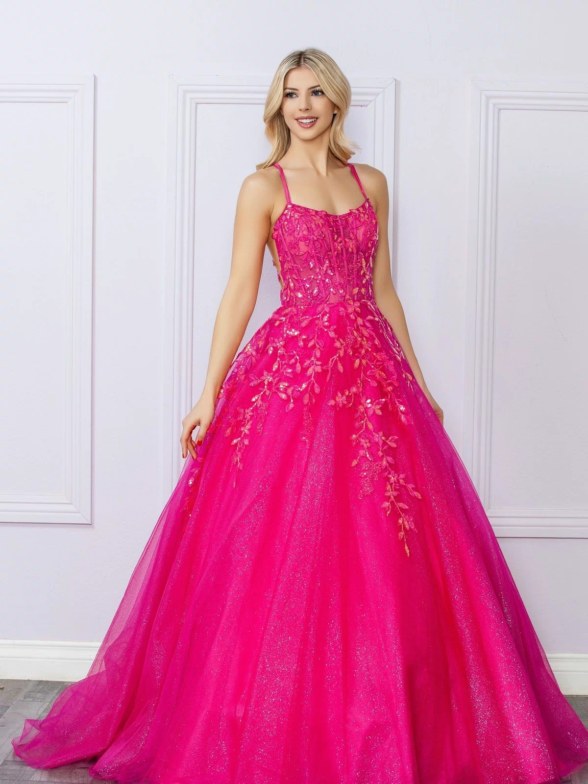 A woman poses indoors against a light-colored, paneled wall wearing the Glitter-Tulle Long Prom Ball Gown H1271, featuring bright pink glitter tulle, floral embellishments, and thin straps.