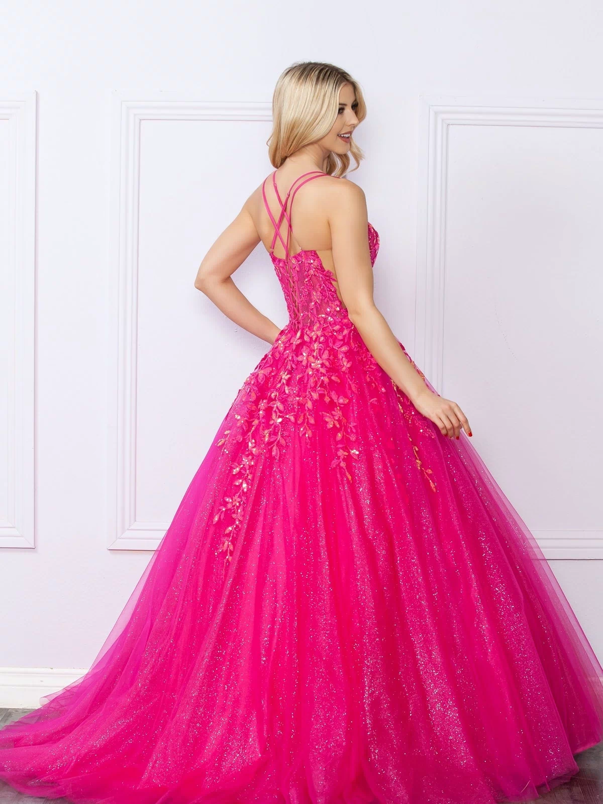 A woman stands with her back to the camera, wearing the Glitter-Tulle Long Prom Ball Gown H1271 in bright pink with floral accents and crisscross straps, its sparkling tulle skirt flowing to the floor against a white wall.