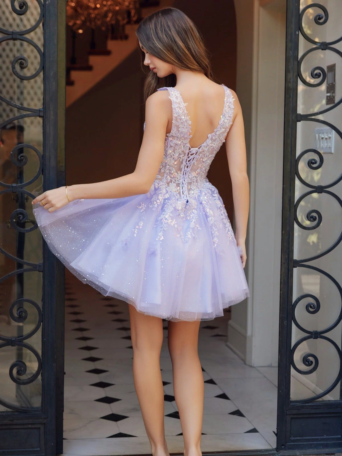 A woman in the Embroidered-Bodice Short A-Line Hoco Dress H846 stands by an open wrought iron gate, holding out one side of her light purple tulle skirt with its embroidered-lace sweetheart bodice.