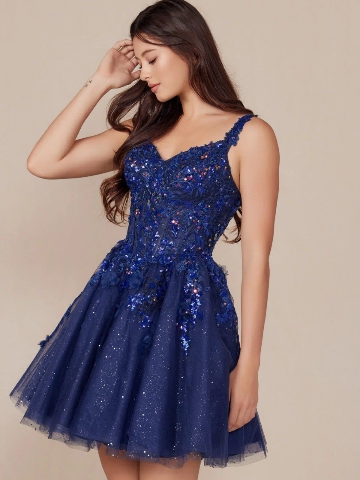 A woman wearing the Embroidered-Bodice Short A-Line Hoco Dress H846, featuring a dark blue sleeveless sequin top and tulle skirt, stands against a beige background, looking down and touching her hair.