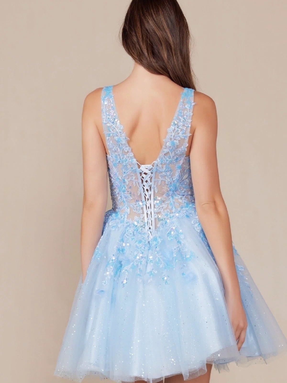 A woman with long brown hair is shown from behind wearing the Embroidered-Bodice Short A-Line Hoco Dress H846, a sleeveless light blue babydoll party dress with a lace-up back, floral appliqué, and an embroidered-lace sweetheart bodice.