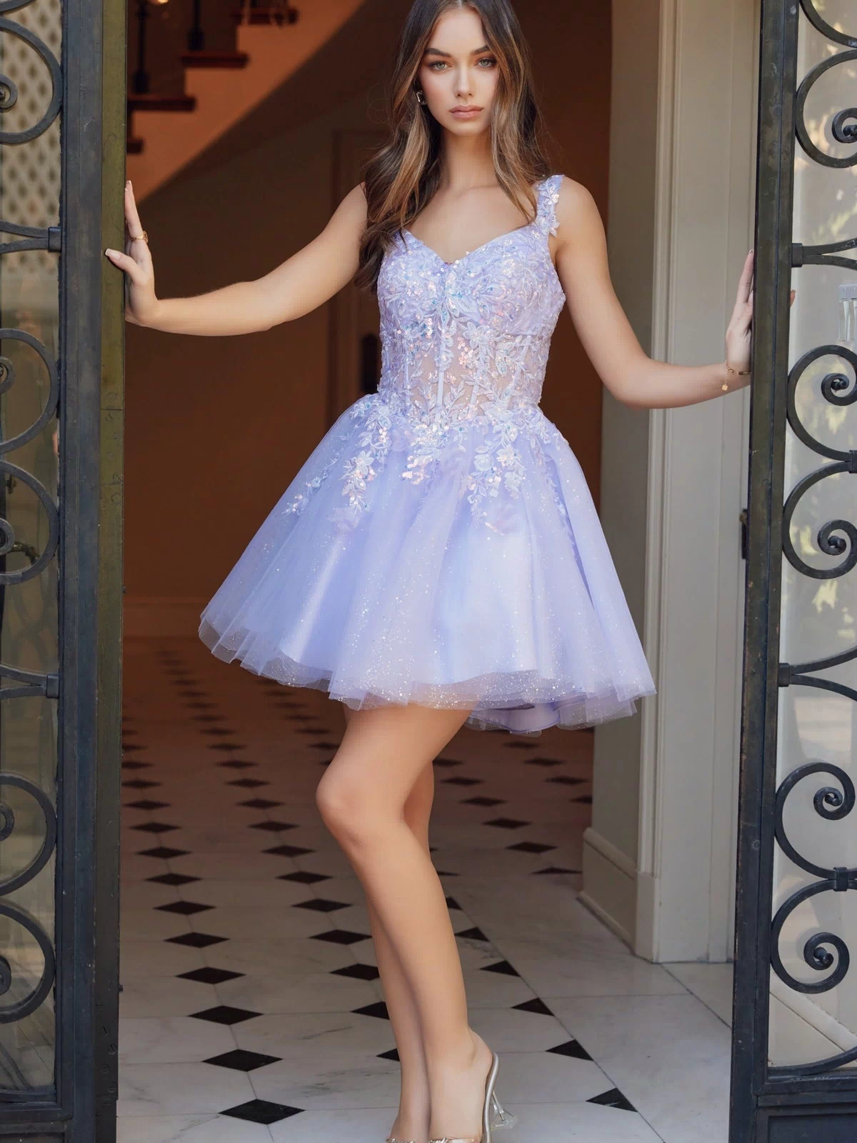 A woman stands in a doorway on a black and white tiled floor wearing the Embroidered-Bodice Short A-Line Hoco Dress H846, featuring floral embellishments and a light purple embroidered-lace sweetheart bodice.
