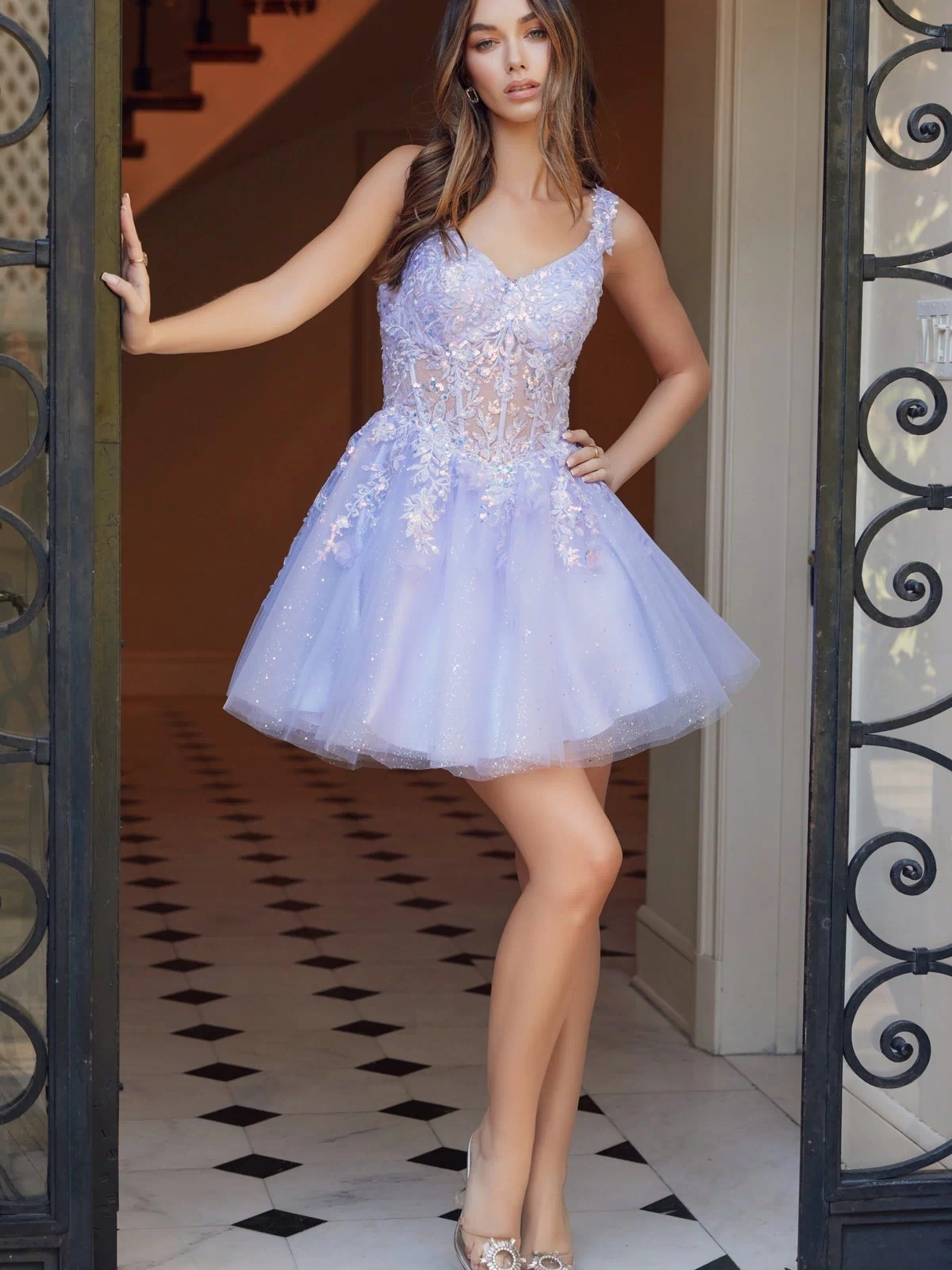 A woman stands in a doorway wearing the Embroidered-Bodice Short A-Line Hoco Dress H846—a light purple, sleeveless babydoll party dress featuring floral details, sparkling accents, and an embroidered-lace sweetheart bodice.