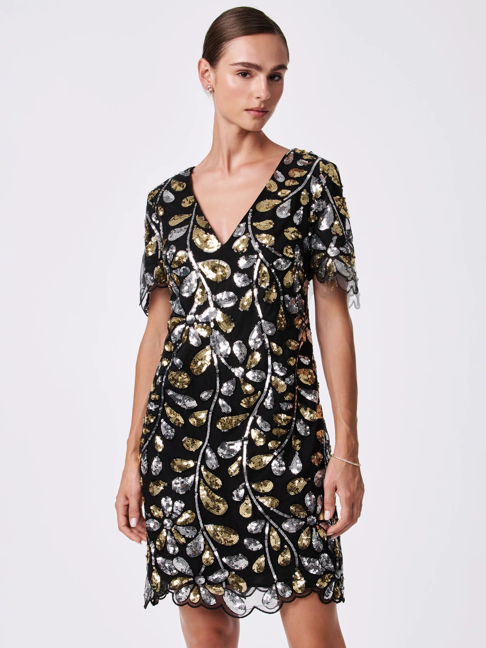 A woman stands against a plain light background wearing the OBELLE 2 DRESS, a black V-neck, short-sleeve dress made from 100% nylon with gold and silver sequin leaf patterns and a scalloped hem.