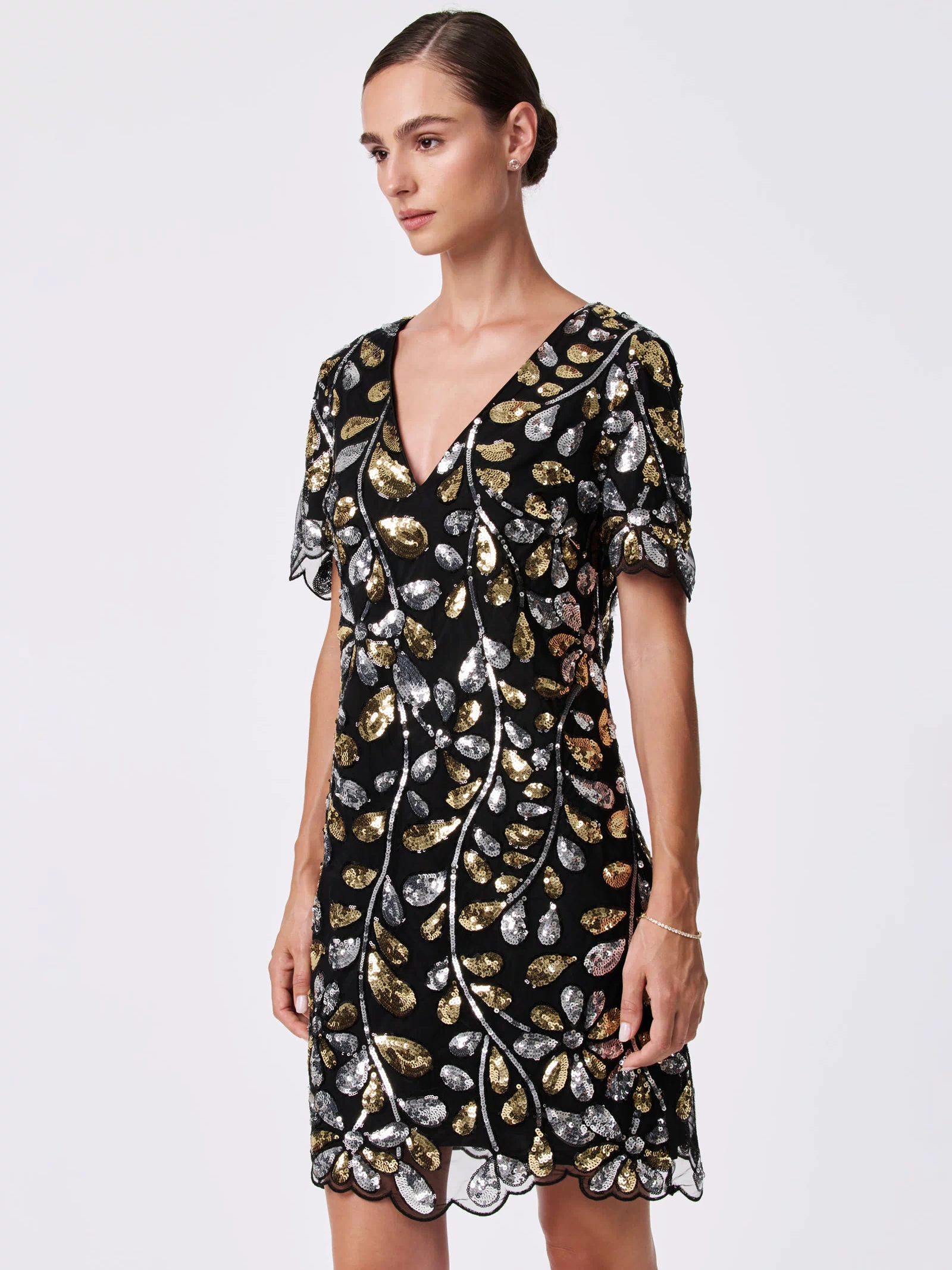 A woman stands against a plain background wearing the OBELLE 2 DRESS, a black 100% nylon dress with gold and silver sequin leaf patterns, short sleeves, and a V-neckline. Her hair is styled in a neat bun.