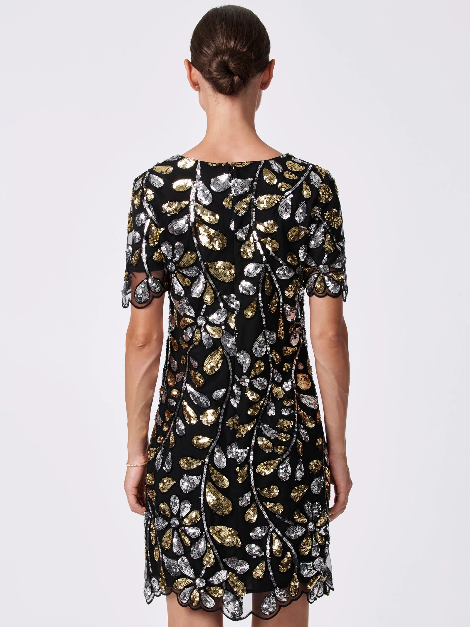 A woman with brown hair in a low bun models the OBELLE 2 DRESS, a short-sleeved black dress made of 100% nylon with gold and silver sequin leaf patterns, facing away against a plain white background.