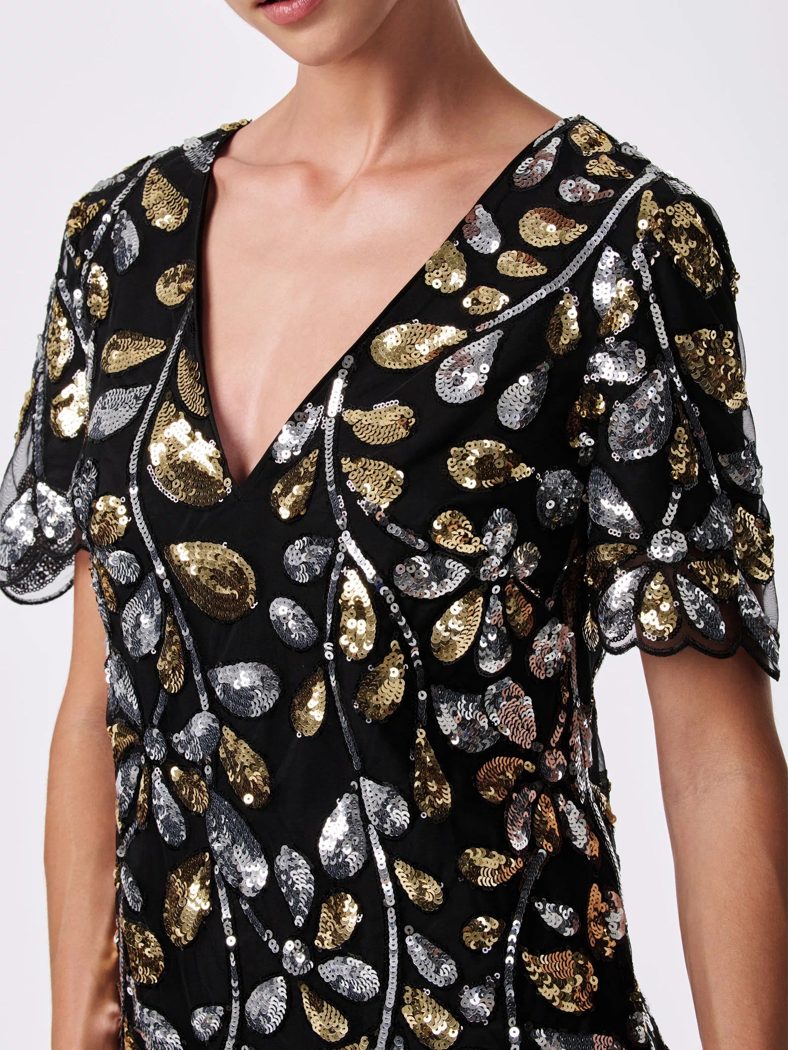 A woman wears the OBELLE 2 DRESS, a black 100% nylon piece with a deep V-neck, short sleeves, and gold and silver sequin leaf patterns. The image is cropped at her upper torso and doesn’t show her full face.