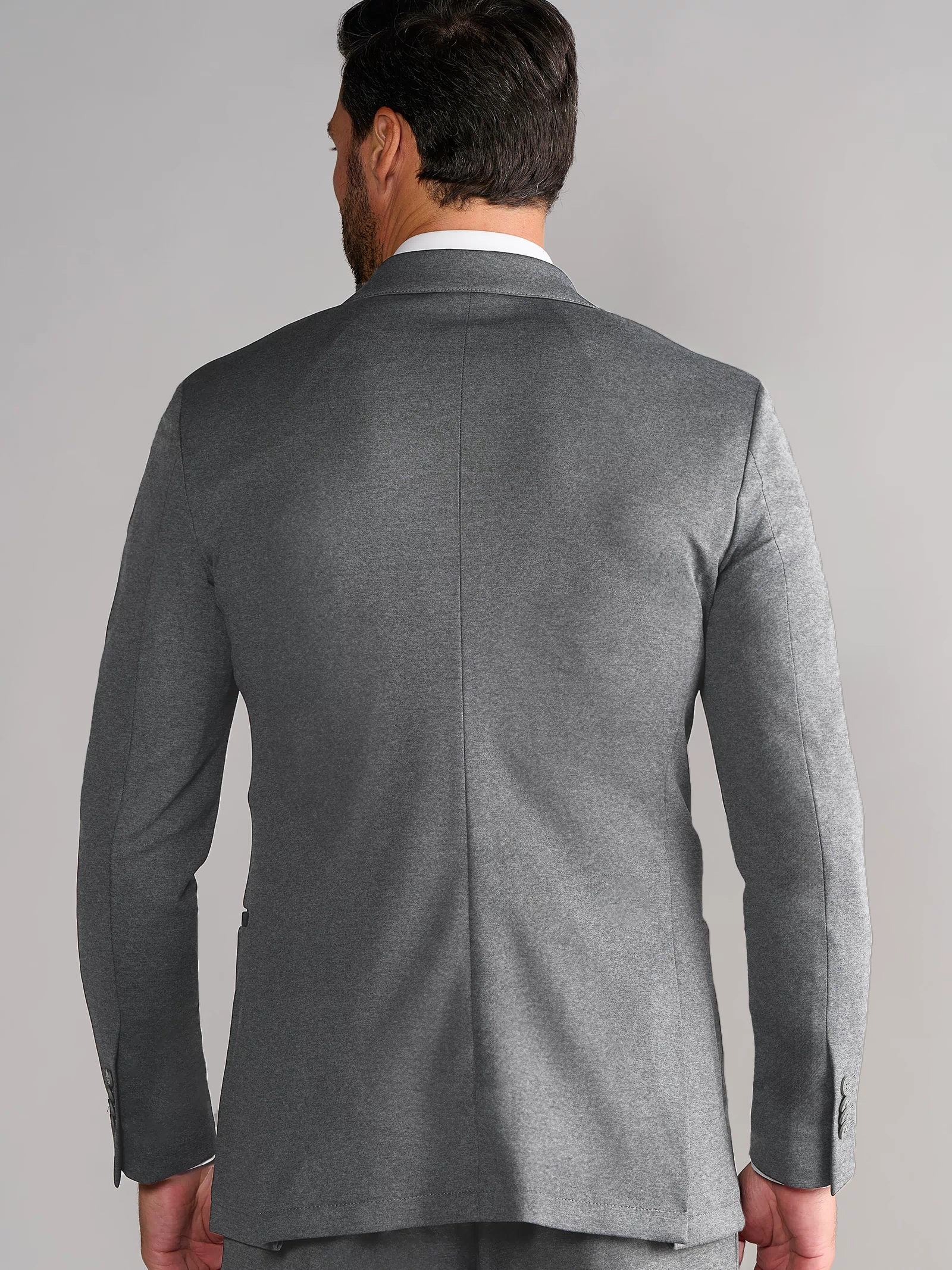 A man with dark hair and a beard is seen from behind wearing Twillory’s Performance Blazer // Heather Light Grey. The plain backdrop highlights the suit’s center back seam and modern stretch design.